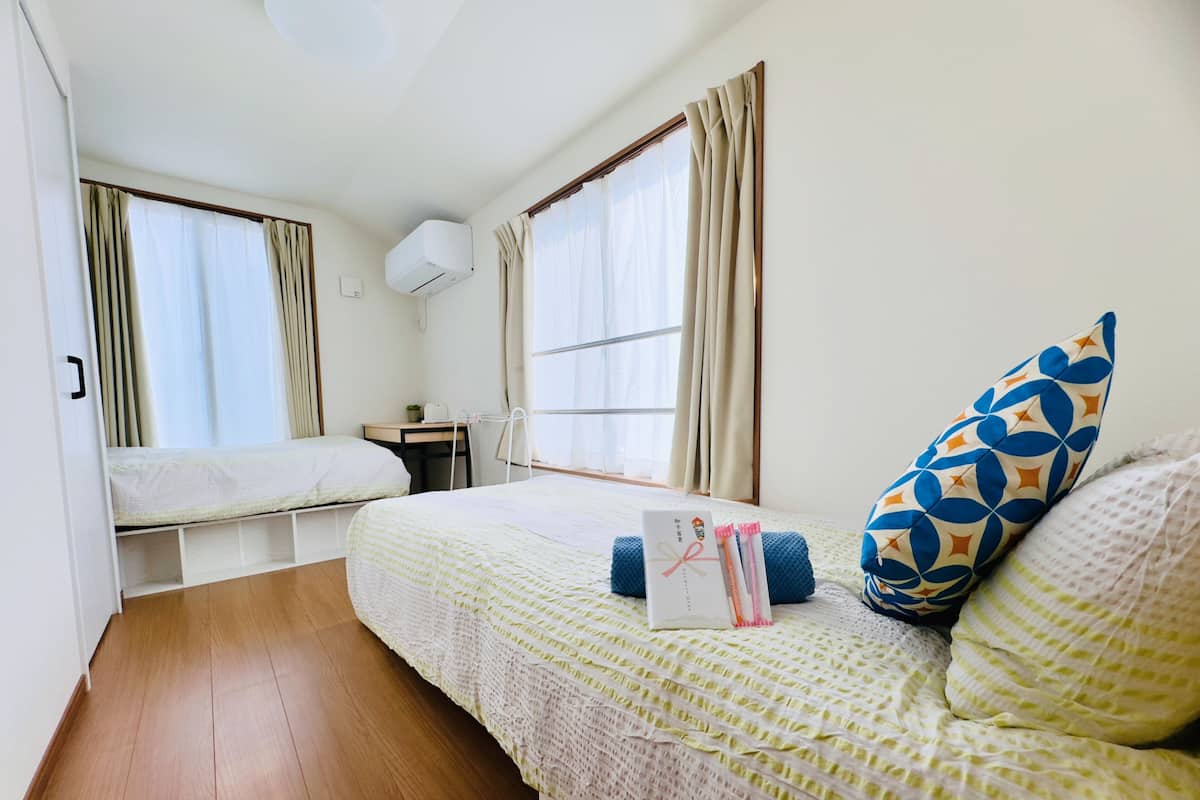 A room with two beds, a desk, a window with curtains, and a wall-mounted air conditioner.