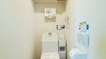 A small, compact bathroom with a toilet, a hand dryer, a paper towel dispenser, and a trash can.