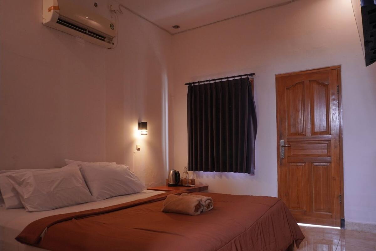A bedroom with a bed, a nightstand, a window with curtains, a wooden door, and an air conditioning unit.