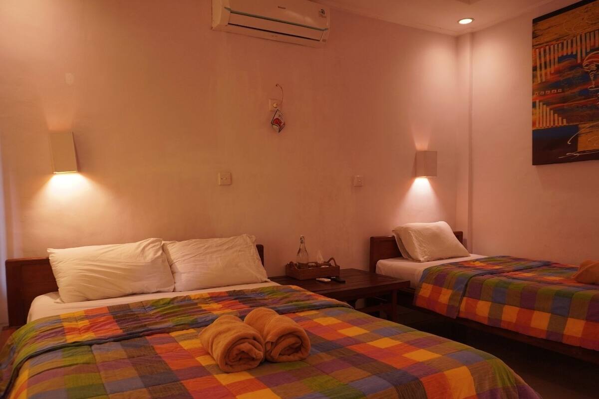 A hotel room with two beds, each with a colorful quilt, a small table with a bottle, and wall-mounted lamps.