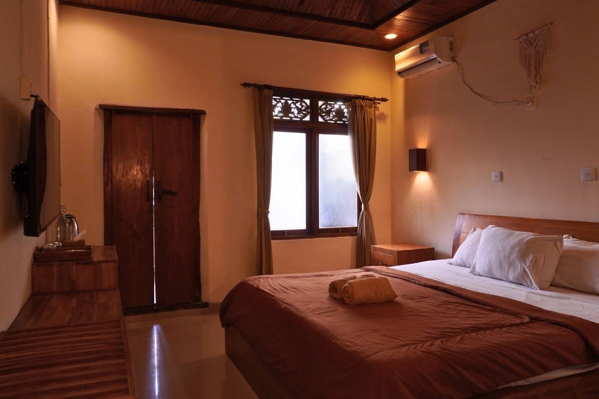A hotel room with a bed, a wooden dresser, a mirror, a window with curtains, and an air conditioning unit.