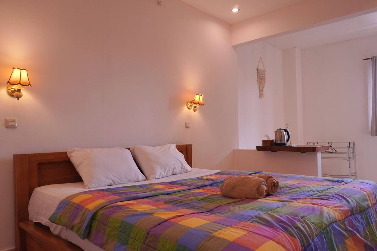 A bedroom with a wooden bed, a colorful quilt, two wall-mounted lamps, a small shelf with a kettle, and a towel on the bed.