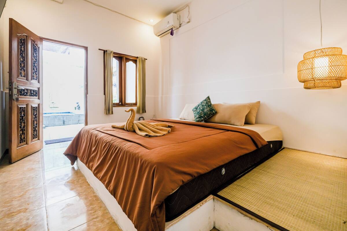 A hotel room with a bed, a window with curtains, an air conditioning unit, and a woven light fixture.