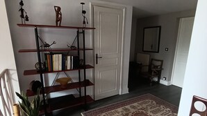 A hallway with a wooden floor, a patterned rug, a bookshelf with various items, a white door, and a mirror on the wall.