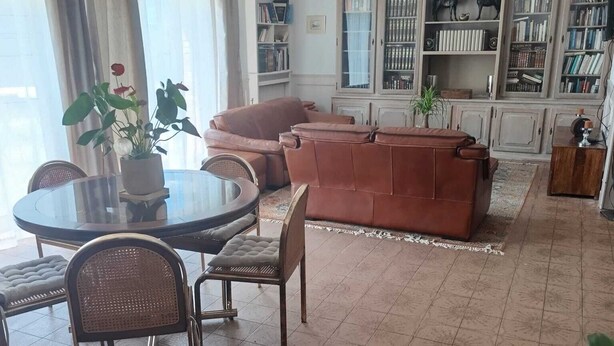 A living room with a brown sofa, a round table, and chairs, a bookshelf, and a dining area with a glass table.