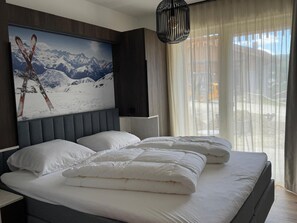 Ein Schlafzimmer mit einem großen Bett, zwei Kissen, einer Kopfteil und einem an der Wand montierten Fernseher, sowie einem Fenster mit Vorhängen.