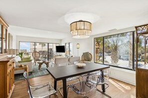 A spacious living area with a large dining table, clear chairs, a modern chandelier, and large windows offering a view of palm trees and a cityscape. - Oceanfront | Private Balcony | Totally Renovated 2nd Floor Amelia Surf and Racquet Condo (Fernandina Beach)
