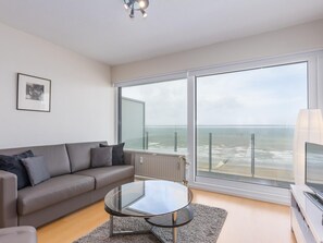 Un salon moderne avec un canapé gris, une table basse en verre et une grande fenêtre donnant sur la mer.