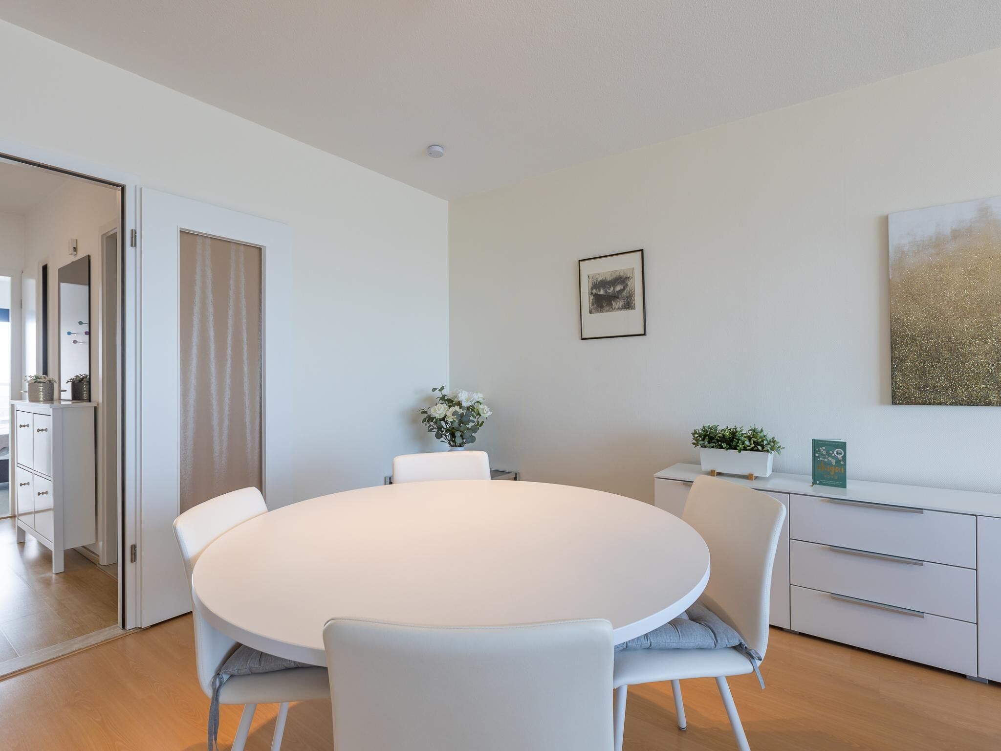 Een moderne eetruimte met een ronde tafel, witte stoelen, een wit kastje en een lijst met een afbeelding aan de muur.