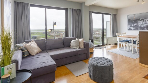 A modern living room with a grey sofa, a round ottoman, a dining table with chairs, and large windows offering a city view. - Apartment on the 8th floor with terrace (Middelkerke)