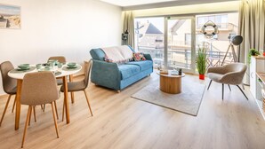 Una sala moderna con un sofá azul, una mesa de comedor y sillas, un balcón con vistas, y una mesita auxiliar con una lámpara.