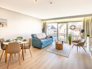 Ein modernes Wohnzimmer mit blauer Couch, Esstisch, Stühlen, einem Balkon mit Blick und einem kleinen Beistelltisch mit Lampe.