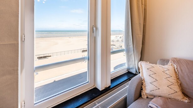 A room with a large window overlooking a beach, a radiator, and a sofa with a patterned cushion. - Tastefully furnished apartment with sea view (Koksijde)