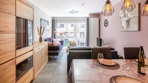 A modern kitchen with wooden cabinets, a dining area with a table set for two, and a living space with a sofa and a TV. - Cozy apartment & terrace, 100 m from sea (Koksijde)