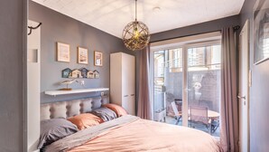 A bedroom with a bed, a hanging light fixture, a window with curtains, and a wall-mounted shelf with decorative items. - Cozy apartment & terrace, 100 m from sea (Koksijde)