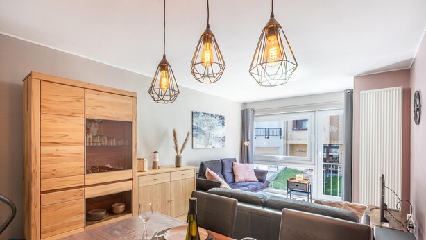 A dining area with a wooden cabinet, a table set for two, and a sofa in the background. - Cozy apartment & terrace, 100 m from sea (Koksijde)