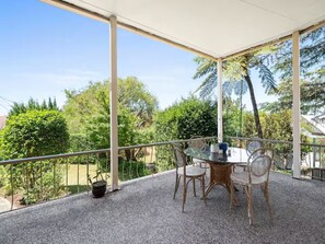 A covered outdoor area with a table and chairs, surrounded by a railing, overlooking a garden with trees and a clear sky. - Spacious Family Home With Iconic Views (Sydney)