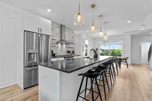 Une cuisine moderne dotée d’appareils électroménagers en acier inoxydable, d’un grand îlot central, de tabourets de bar noirs et de luminaires suspendus.
