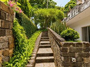 Eine steinerne Treppe führt zu einem Gebäude, umgeben von üppigem Grün und einer blühenden Pflanze in Rosa.