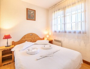 A neatly made bed with pillows and a blanket, a bedside table with a lamp, a framed picture on the wall, and a window with sheer curtains. - Duplex for 12 Near Lake Annecy (Giez)