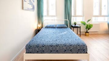 Een bed met een bedspreide, een nachtkastje met een lamp, een kamerplant en een raam met gordijnen.