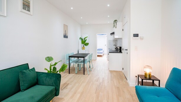 A modern living room with a green sofa, a blue sofa, a dining table with chairs, a kitchen area, and a hallway leading to another room. - Cosy Apartment in Central Antwerp (Antwerp)