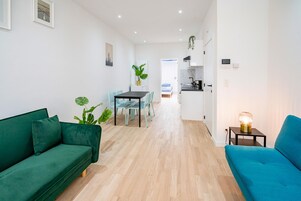 Uma sala moderna com um sofá verde, um sofá azul, uma mesa de jantar com cadeiras, uma cozinha e um corredor que dá para outro cômodo.