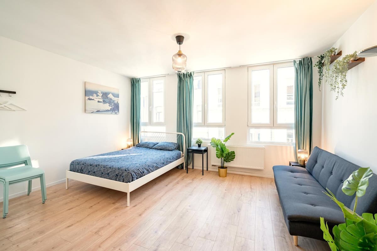 A bedroom with a bed, a sofa, a chair, a small table, and a potted plant.