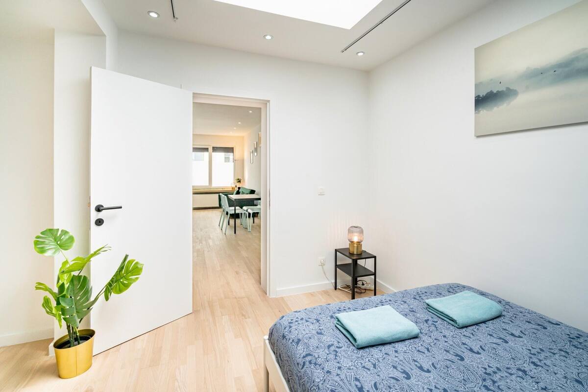 A bedroom with a bed, a bedside table, a potted plant, and a view into the dining area.
