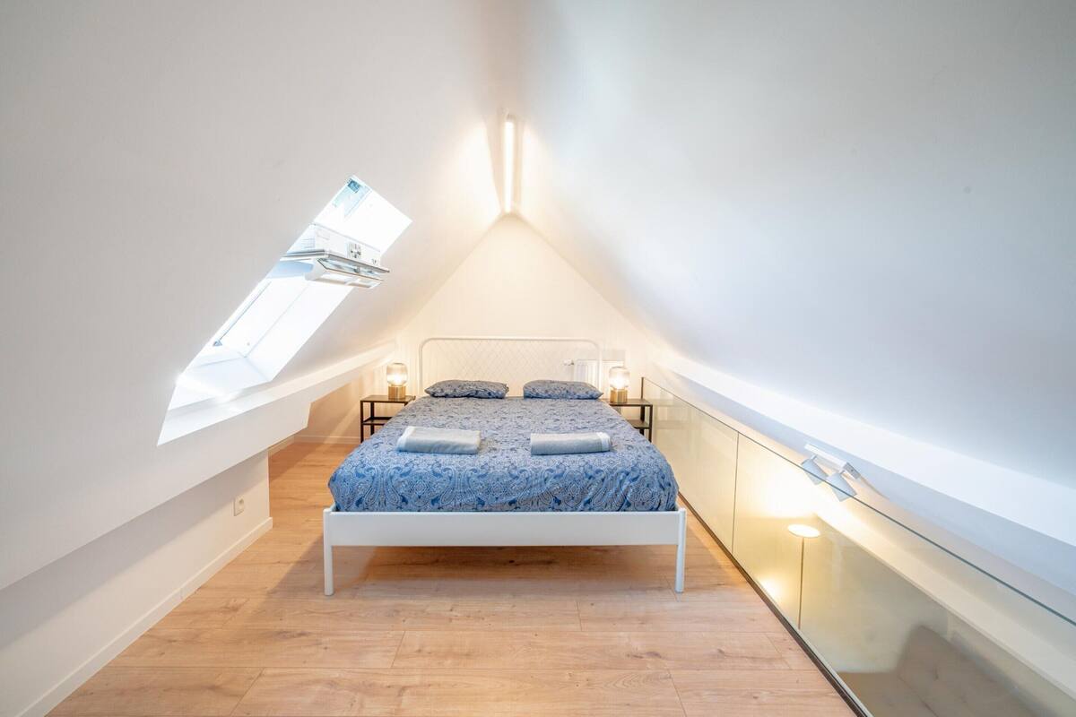 A bedroom with a sloped ceiling, a skylight, a bed with a patterned bedspread, bedside lamps, and a glass railing along the wall.