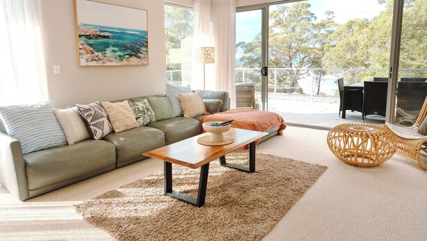 Ein Wohnzimmer mit einer Couch, einem Couchtisch, einem Korbstuhl und Blick ins Freie durch große Glasfenster.