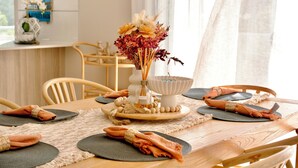 Ein Esstisch gedeckt mit Tischsets, gefalteten Servietten und einer Blumenarrangement in der Mitte.