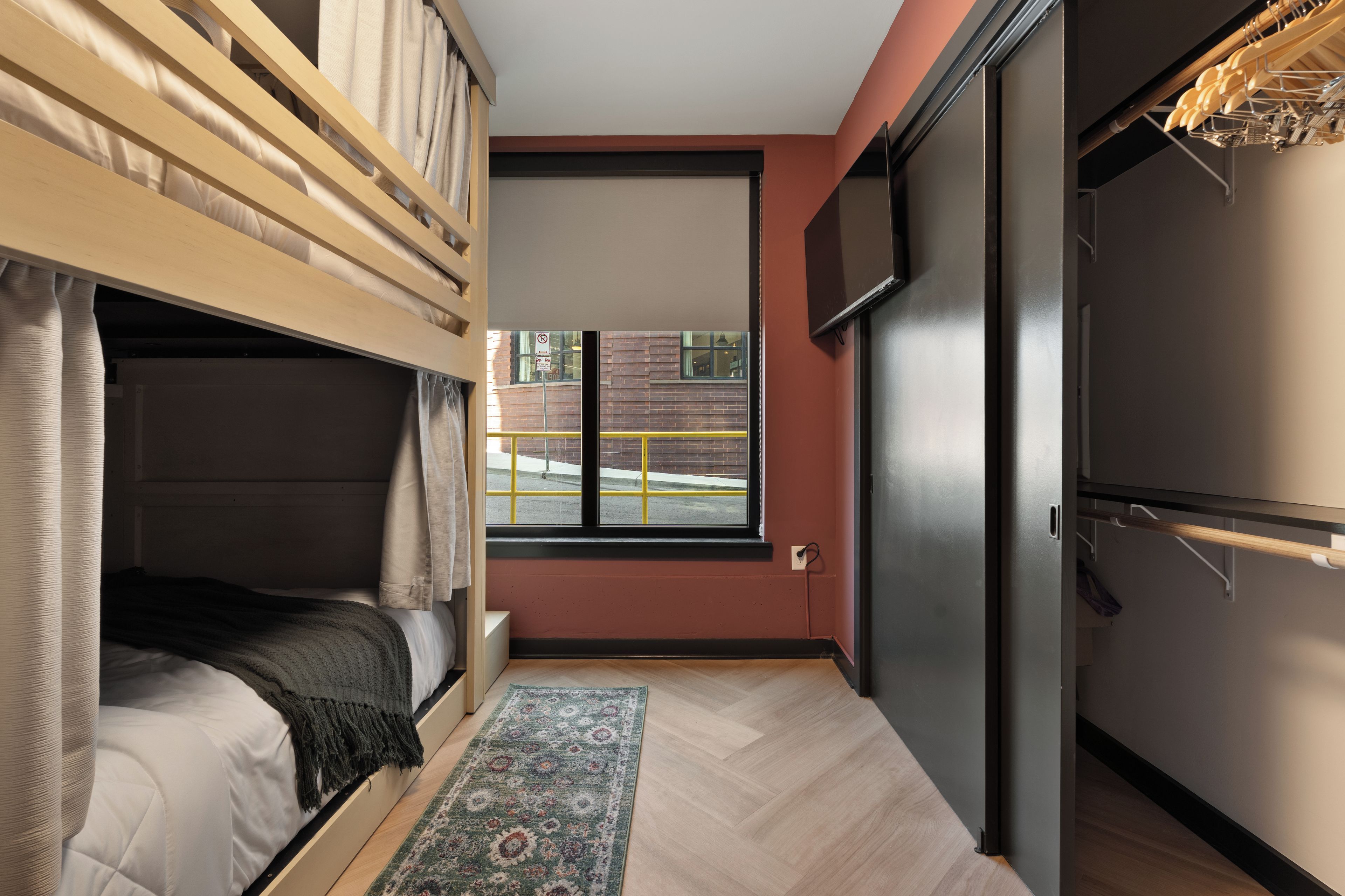 A compact room with a bunk bed, a small window with a view of a building, a flat-screen TV mounted on the wall, and a wooden door.