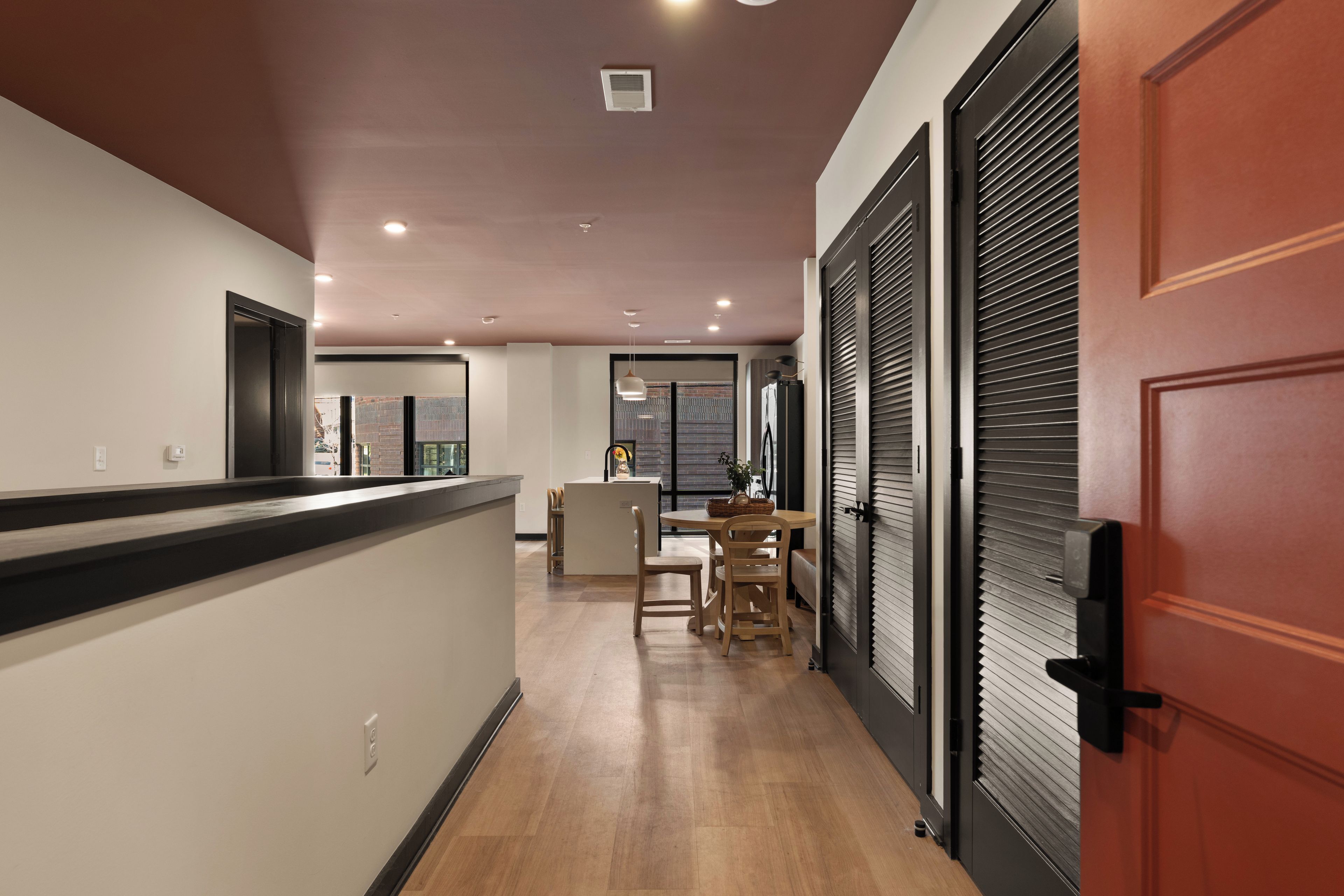 Un pasillo con suelo de madera, un mostrador de recepción, una zona de comedor con sillas y una mesa, y una puerta cerrada con manija.