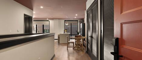 Un pasillo con suelo de madera, un mostrador de recepción, una zona de comedor con sillas y una mesa, y una puerta cerrada con manija.