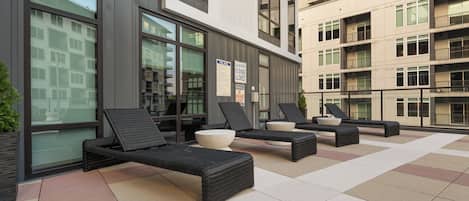 Zona de estar exterior con sillones reclinables y una mesa junto a la piscina, adyacente a un edificio con grandes ventanales y un letrero con las normas de la piscina.
