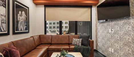 Ein modernes Wohnzimmer mit einem braunen Ledersofa, einem Couchtisch mit Pflanze und Büchern sowie einem großen Fenster mit Blick auf Gebäude.
