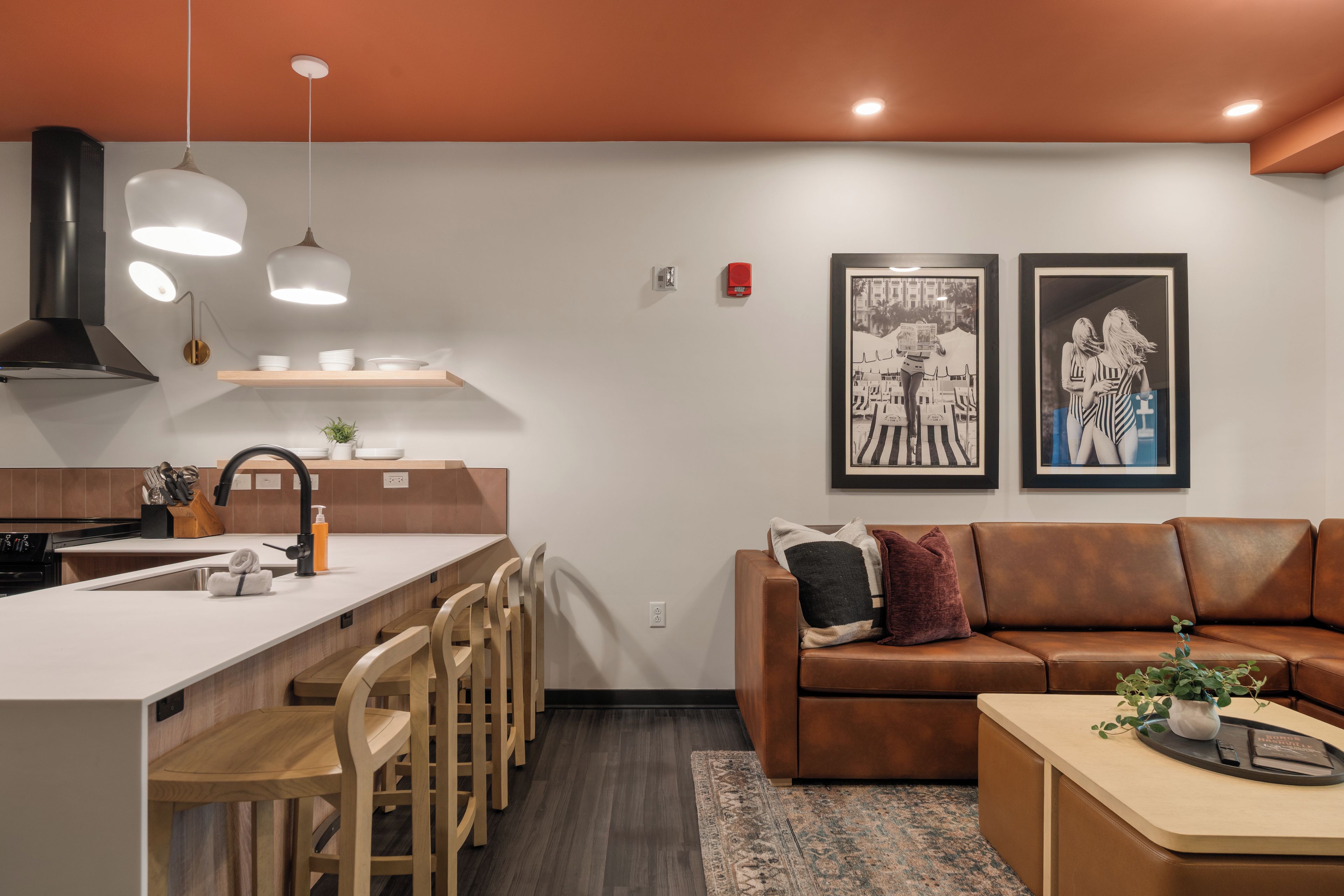 A modern kitchen with a central island, bar stools, and a dining area. There is a brown leather sofa and a small table with a tray and a plant.