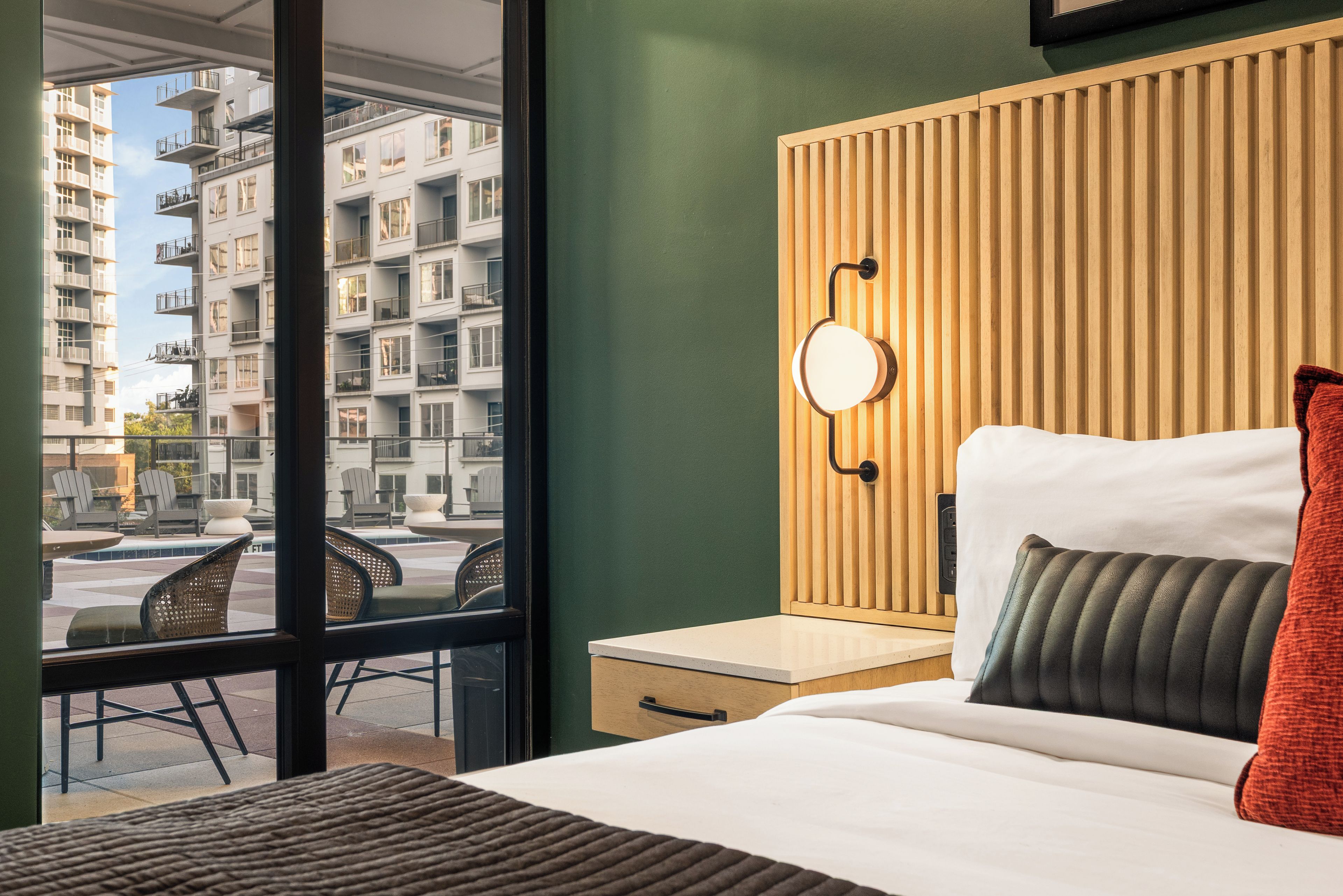 A hotel room with a large window, a bed with a white headboard, a nightstand, and a wall-mounted light fixture.