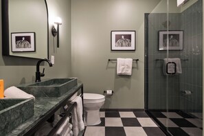 A bathroom with a black and white checkered floor, a white toilet, a glass shower enclosure, a marble sink, and two framed pictures on the wall. - Superior Suite - Unit 401 (Nashville)