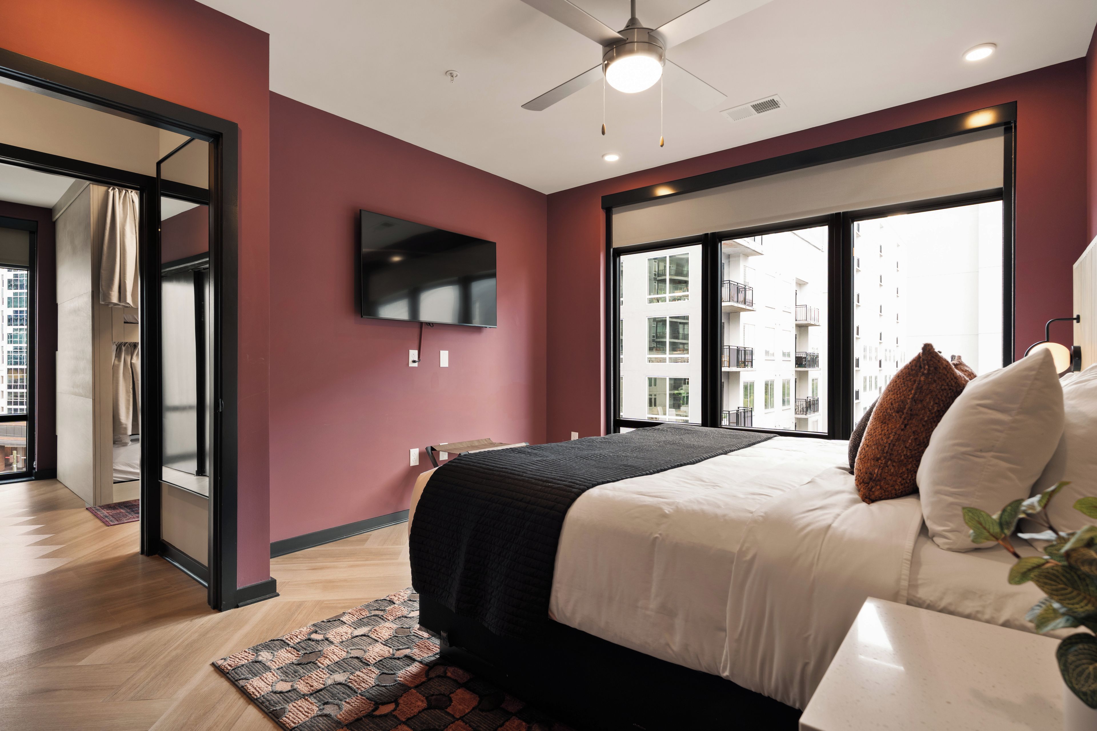 Un dormitorio moderno con cama, televisor de pantalla plana, ventilador de techo y ventanas amplias.