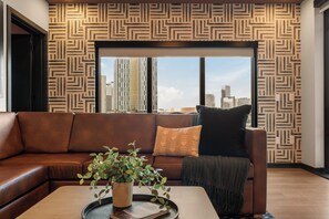 Una sala de estar moderna con una pared estampada, un sofá de cuero, una mesa de centro con una planta y vistas a un paisaje urbano a través de ventanas amplias.