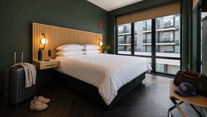 A hotel room with a bed, bedside table, lamp, suitcase, and a view of the building outside.