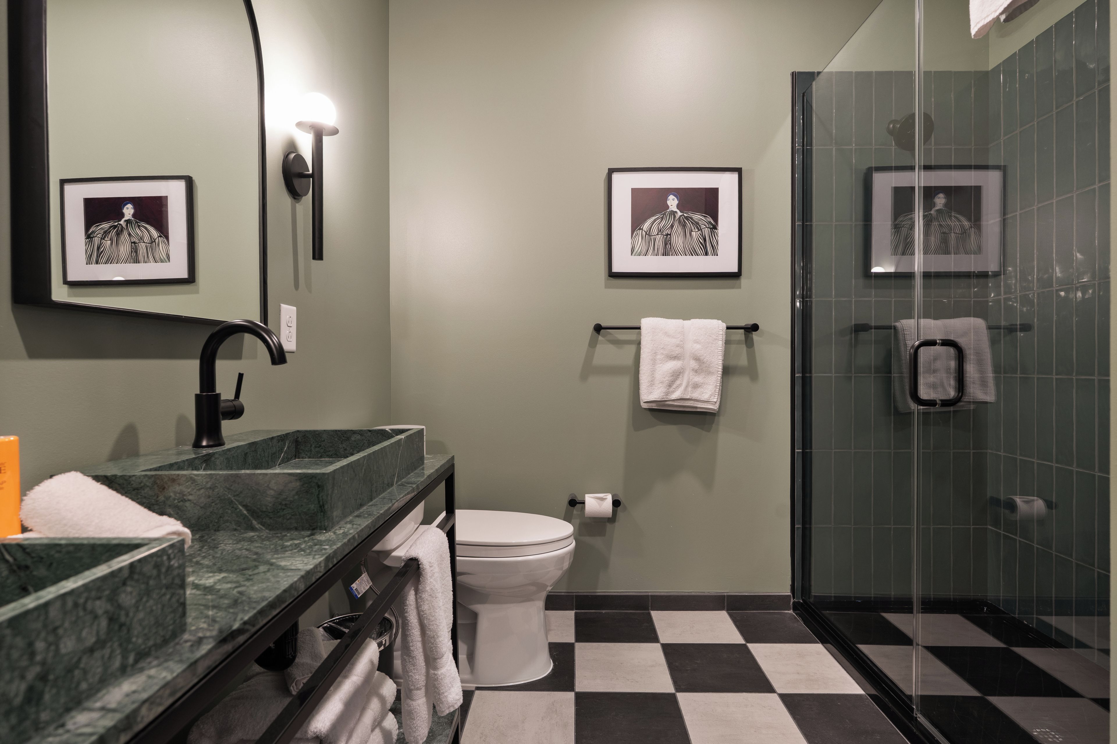 Ein Badezimmer mit schwarz-weiß kariertem Boden, einer weißen Toilette, einer Duschkabine aus Glas, einem мраморном Waschbecken und zwei gerahmten Bildern an der Wand.