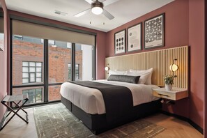 Un dormitorio moderno con una cama grande, una mesita de noche, una lámpara de pared y vista a un edificio de ladrillo a través de un ventanal amplio.