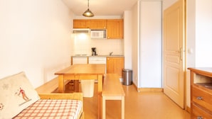 A compact room with a bed, a wooden dining table, a kitchen area with wooden cabinets and a microwave, and a door. - Cosy Alpine Studio With Wellness Access (Saint-Sorlin-d'Arves)