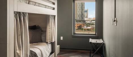 A room with a bunk bed, a window with a city view, a red patterned rug, and a ceiling light.