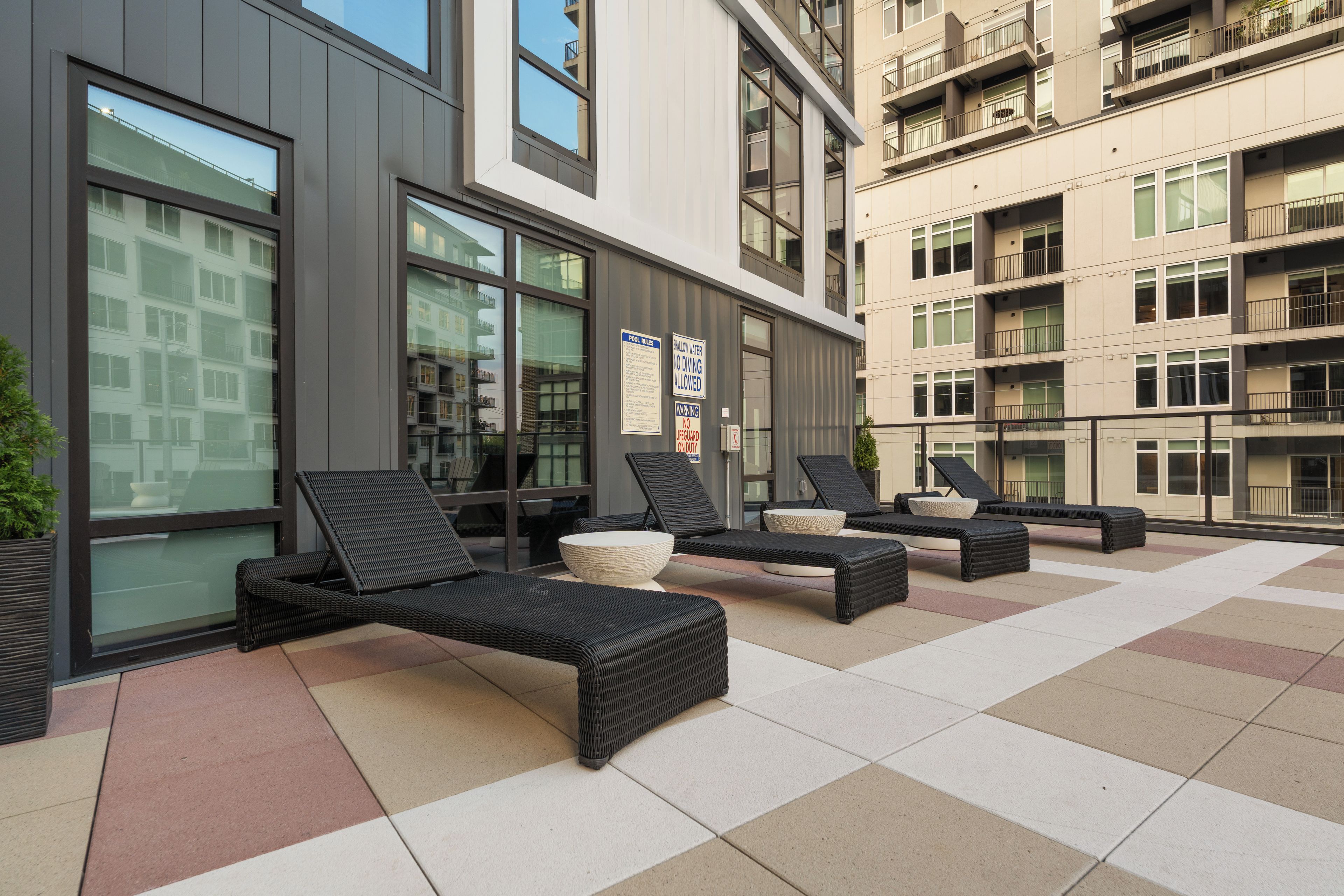An outdoor seating area with lounge chairs and a poolside table, adjacent to a building with large windows and a sign indicating pool rules.