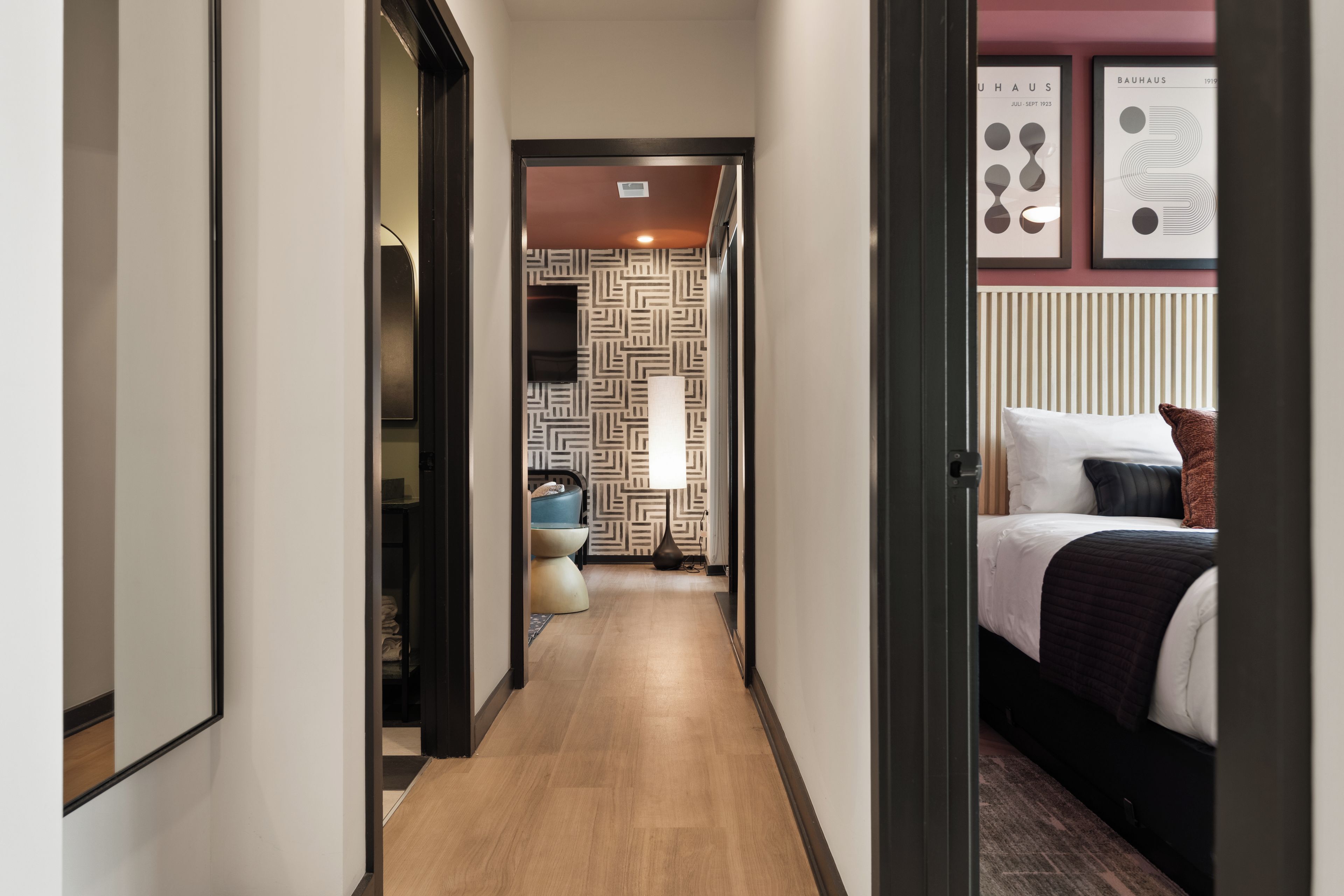 A hotel hallway with a wooden floor, a bed with a black and white bedspread, and a wall with framed artwork.