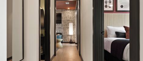 A hotel hallway with a wooden floor, a bed with a black and white bedspread, and a wall with framed artwork.